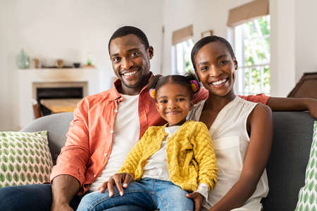 Portrait of smiling african american family relaxing on couch with copy space. Happy mature black man and beautiful woman sitting on sofa at home with daughter looking at camera. Portrait of happy mother and father with little cute girl sitting on couch cuddling.の写真素材
