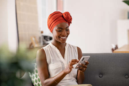 Smiling middle aged african woman with traditional head turban sitting on couch at home using smartphone. Beautiful african american woman with typical headscarf scrolling through internet on cellphone. Happy mid adult lady sitting on sofa browsing mobile phone.の写真素材