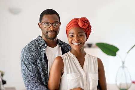 Portrait of cheerful middle aged couple embracing at home. Romantic mid adult black man hugging beautiful wife with traditional red turban while smiling and looking at camera. Happy mature african couple posing.の写真素材