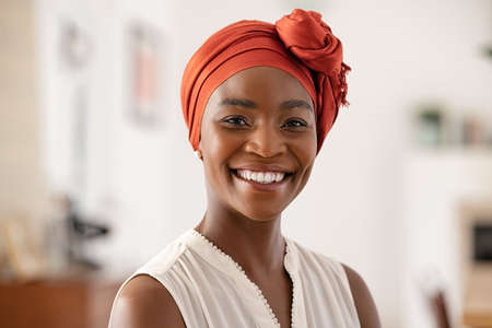 Portrait of smiling middle aged African american woman with headscarf at home. Cheerful mid adult black woman with turban looking at camera at home. Happy mid mature lady wearing traditional african scarf on head.の写真素材
