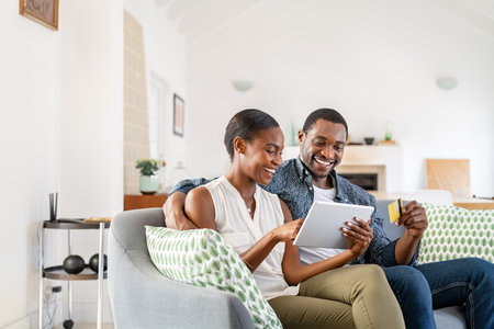 Happy african american wife doing shopping online while using digital tablet and credit card at home with husband. Middle aged black couple relaxing at home while making an online purchase using debit card on digital tablet. Mid couple using bank card to book holidays.の写真素材