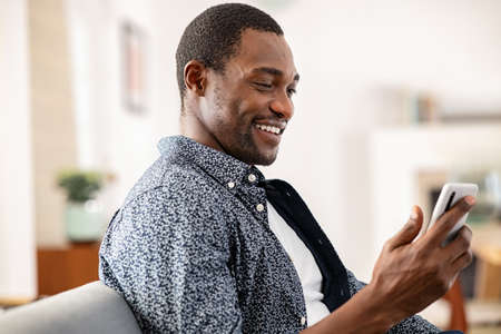 Happy mid adult man relaxing on couch at home while using smartphone. Handsome black man reading text message on cellphone. Middle aged smiling african guy resting at home and doing a video call with mobile phone.の写真素材