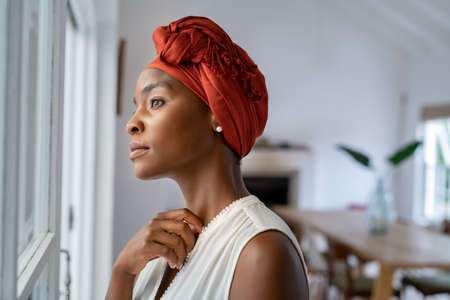 Beautiful pensive woman wearing headscarf looking outside window in contemplation. Mature black woman wearing a traditional turban and standing near window at home while thinking. Worried african mature woman with cancer at home.の写真素材