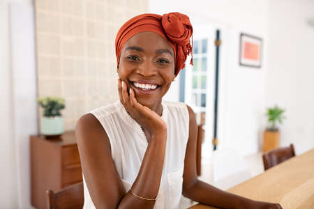 Portrait of smiling middle aged woman wearing african turban. Happy mid african american woman with hand on chin wearing traditional turban and looking at camera. Beautiful black mature woman stay at home.の写真素材