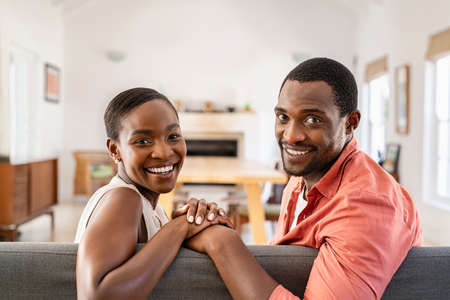 Portrait of mature black couple looking at camera while sitting on sofa. Happy african american couple relaxing at home. Middle aged man holding hands of his beautiful ethnic wife while smiling together on couch at home.の写真素材