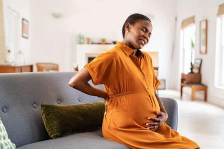 Mature pregnant woman having a backache while relaxing on couch at home. Unhappy black expecting woman suffering from lower back pain sitting on sofa with copy space. Tired middle aged african pregnant woman suffer from lower back pain on last month of pregnancy.の写真素材