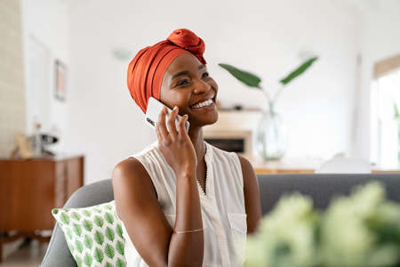 Smiling middle aged woman sitting on couch at home talking over smartphone. Happy black woman with traditional head turban using mobile phone for conversation. Relaxing mid african american lady sitting on sofa at home while using cellphone.の写真素材