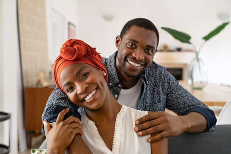 Portrait of mid adult happy african american couple hugging at home while sitting on couch in living room. Cheerful middle aged black man embracing his smiling wife from behind. Portrait of mature man with his beautiful woman wearing traditional turban relaxing at home and looking at camera.の写真素材