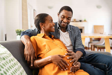 Happy mature loving husband and black pregnant woman embracing belly at home. Smiling middle aged african american man hugging pregnant woman sitting on couch holding tummy. Beautiful mid adult couple expecting baby and dreaming on sofa.の写真素材