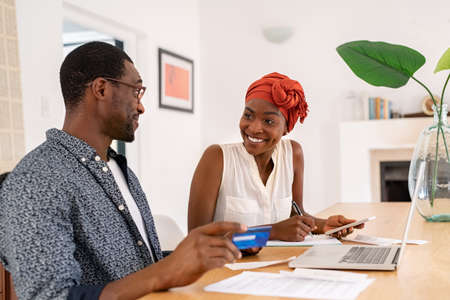 Mature smiling couple sitting and managing expenses at home. Happy mid black man and woman with traditional turban paying bills and managing budget. Middle aged african american couple checking accountancy and bills while holding credit card.の写真素材