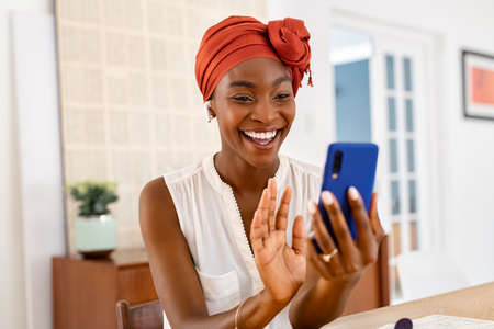 Portrait of smiling african american woman waving hand while on video call using smartphone. Happy black woman in conversation using cellphone at home and wearing traditional turban. Beautiful middle aged lady using mobile phone during video conference at home.の写真素材