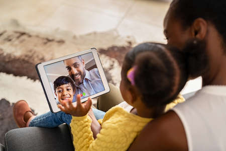 Black mother and daughter using digital tablet while sitting on couch doing a video call with middle eastern father and little son. African american woman and lovely daughter on video call with family using digital tablet at home. Cute little girl doing video conference with her schoolmate during the lockdown.の写真素材