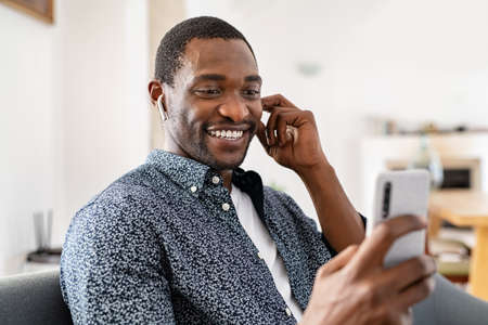 Smiling mid adult african man relaxing on couch at home while using smartphone to do a video conference. Middle aged smiling man resting at home and using wireless earphones while talking on a video conference with friend. Businessman using video chat while working from home.の写真素材