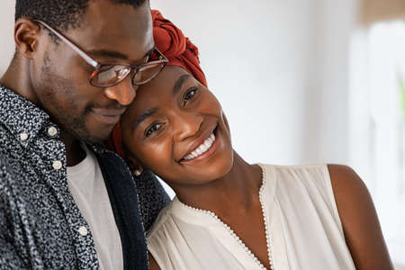 Portrait of happy african american woman relaxing on man shoulder wearing traditional turban. Smiling mid adult couple in love hugging each other at home. Beautiful mature woman wearing red headscarf while embracing husband at home and looking at camera.の写真素材