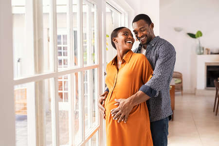 Lovely mid black man hugging his pregnant wife from behind standing near window at home. Happy middle aged black husband embracing pregnant woman while waiting for baby. African american mature couple hugging and looking at each other.の写真素材