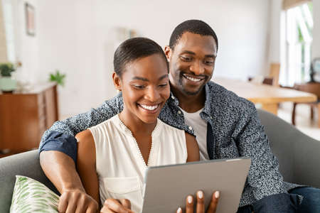Cheerful black couple sitting on couch and watching movie on digital tablet at home. Mature black husband and beautiful wife using digital tablet to do a video call while sitting on sofa. Middle aged man and african woman relaxing on couch in living room.の写真素材