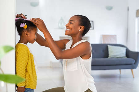 Happy and black mature mother measuring daughter height on wall with marker at home. Beautiful middle aged woman tracking increase in height of little girl. Cheerful mid african american woman noting growth with book and pencil of daughter on home wall.の写真素材
