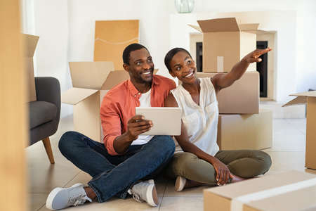 Happy middle aged couple planning decoration after moving home sitting on the floor with cardboards around. Excited african american couple sitting and planning new home furnishings with a digital tablet at home. Mid adult man and beautiful woman moving house together and planning online with a digital tablet.の写真素材