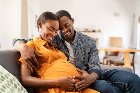 Happy african american husband and pregnant woman hugging belly at home. Smiling black man hugging happy pregnant wife sitting on sofa and holding tummy. Mature loving couple expecting their first baby.の写真素材