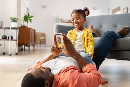 Cute little african american girl sitting on father's stomach and playing with him. Mature black father and daughter enjoying at home by clicking pictures in mobile phone. Lovely dad taking picture of her little cute girl with smart phone at home.の写真素材