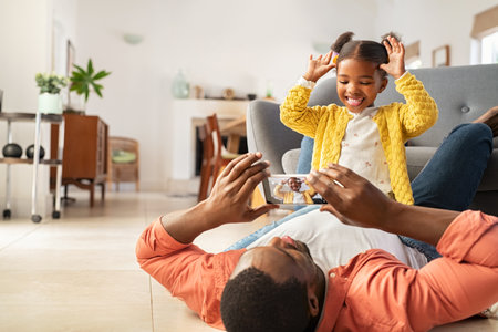 Cute little african american girl making funny face sitting on father's stomach taking photo on smartphone. Middle aged black father and daughter enjoying at home by clicking pictures in mobile phone. Little girl with father having family time and playing together at home.の写真素材
