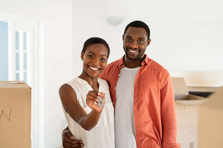 Cheerful mid african wife holding new house keys while standing with husband. Portrait of smiling man embracing beautiful wife holding keys of new house. Happy mature black couple relocating and looking at camera.の写真素材