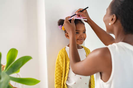 African middle aged mother measuring daughter height and marking it on wall at home. Happy black mother measuring cute little girl height at home using book and pen. Cheerful mature woman measuring her daughter's progression against white wall.の写真素材