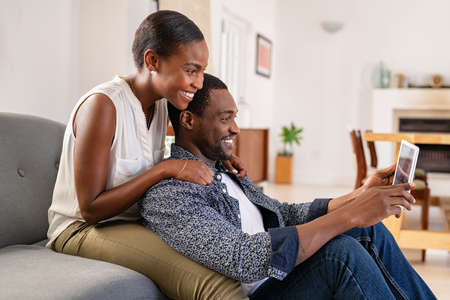 Middle aged black couple using digital tablet while sitting on couch to watch movie on streaming app. Happy smiling black woman embracing boyfriend from behind while watching video on digital tablet at home. African american woman doing video call with husband on digital tablet at home.の写真素材