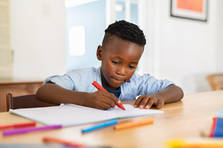 Little black boy sitting on table and painting with colored marker on book. Cute african american child drawing in the living room at home. Schoolboy colouring in book with red marker at home.の写真素材