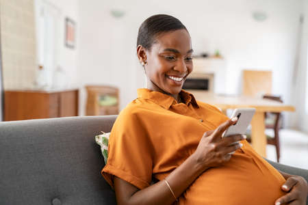 Beautiful mature pregnant woman watching funny video on smartphone. Happy black pregnant woman using mobile while touching tummy at home with copy space. Cheerful middle aged expecting african american woman using cellphone while relaxing on couch at home.の写真素材