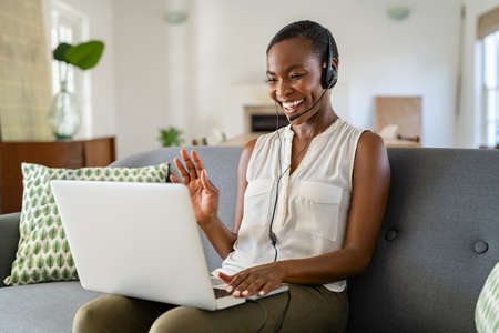 Smiling middle aged african american woman with headphone and mic waving hand during video call on laptop. Cheerful black mid adult woman enjoying conference on laptop while sitting on couch at home. Happy mature lady using computer for video call while working from home.の写真素材