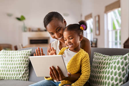Smiling ethnic mother and funny daughter on video call using digital tablet at home. African american mature mother and little girl using digital tablet while making video call and waving hello sitting on sofa. Happy black woman and her cute little kid sitting on sofa and doing video conference with grandparents.の写真素材