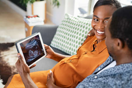 Black mature pregnant couple watching ultrasound picture on digital tablet. Smiling black pregnant woman looking at husband while feeling happy and excited on seeing baby ultrasound on digital tablet. Pregnant mature woman looking her baby on the tablet with her husband while sitting on couch at home.の写真素材