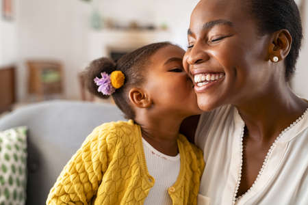 Close up of beautiful daughter kissing mother on cheek at home. African american little girl giving kiss to happy mother. Lovely black female child kissing cheerful and proud woman on cheek with copy space for mother's day.の写真素材