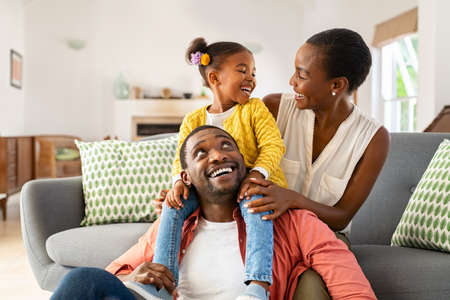 Cheerful little girl sitting on father shoulder while playing with mother at home. Happy black  family enjoying weekend at home. Cute little daughter sitting on fathers shoulder and play with her mom.の写真素材