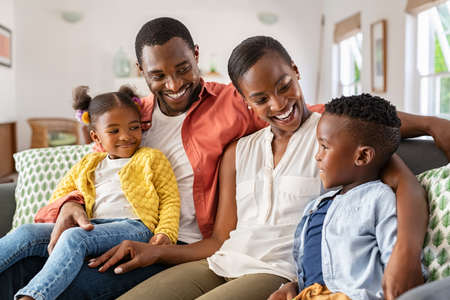Smiling african american family relaxing on couch and talking to each other. Happy black father and mother sitting on sofa at home with their daughter and son. Playful ethnic family with two little kids sit on couch cuddling.の写真素材