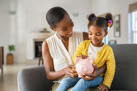 Smiling mature african american mother helping daughter sitting on lap putting money in piggy bank. Cute little black girl saving money by adding a coin in piggy bank with mother at home. Happy daughter sitting on momâs laps and putting coins into piggybank.の写真素材
