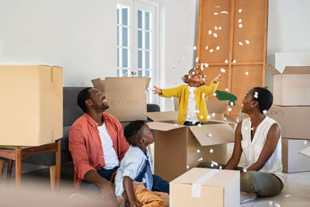 Happy black little girl playing with polystyrene foam chips while standing inside a large box. Happy african american family enjoying new home. Mature parents with son and funny daughter having fun during moving house.の写真素材