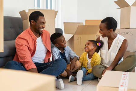 Happy family with two children having fun at new home. Middle-aged black parents with son and daughter in their new house with cardboard boxes after relocation. Smiling little boy and girl sitting on floor with mother and dad.の写真素材
