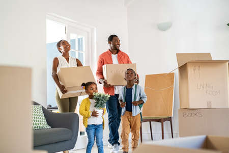 Beautiful african american family with two children carrying boxes in a new home. Cheerful mature mother and mid adult father holding boxes while entering new home with son and daughter. Happy son and daughter helping parents relocating in new house with copy space.の写真素材