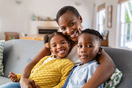 Portrait of mature happy mature mother with son and daughter looking at camera. Middle aged black woman with two children sitting on couch at home while smiling. Beautiful ethnic family relaxing at home on sofa.の写真素材