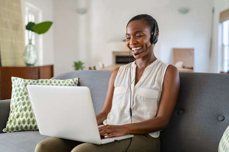 Successful middle aged african american woman with headphone and mic talking on video call using laptop. Cheerful mid black woman enjoy video call on laptop while sitting on couch at home. Happy smiling lady using computer for video conference while working from home.の写真素材