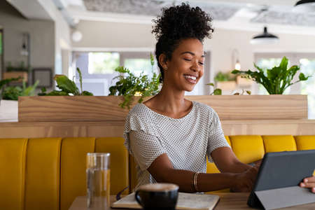 Young black woman entrepreneur working on digital tablet in cafe. Happy smiling woman sitting in cafeteria using digital tablet. Successful black university student with curly hair sitting on modern coffee shop while studying on laptop and doing breakfast.の写真素材