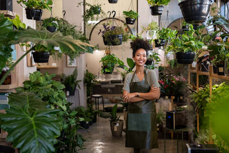 Portrait of african american woman with crossed arms wearing apron standing in botanical store. Smiling young woman in botany store standing between plants looking at camera. Happy small business owner working at flower shop standing surrounded by plants.の写真素材