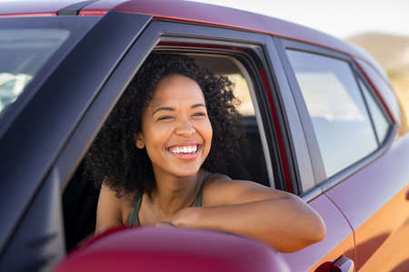 Beautiful african american woman looking outside car during summer holiday. Young attractive black woman laughing and enjoying road trip on a summer day. Happy girl driving a red car with big grin.の写真素材