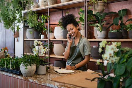 Smiling woman seller in botanical shop talking on smartphone while writing in diary. African american botanist writing client order in notebook during conversation on mobile phone in store. Young black small business owner wearing apron receive client order for plant and delivery at greenhouse.の写真素材