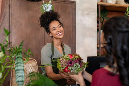 Portrait of smiling black botanist selling a bouquet of fresh flowers to a customer in a botany shop. Happy african woman entrepreneur standing behind counter wearing apron and selling bunch of fresh flowers to client. Young latin woman buying plant in flower shop.の写真素材