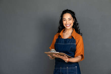 Portrait of smiling young woman in apron holding clipboard and looking at camera. Beautiful latin waitress waiting for customers isolated on grey wall. Successful small business owner standing on grey background with copy space.の写真素材
