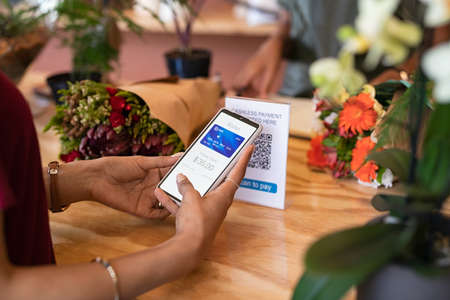 Close up of woman hands holding smartphone scanning QR code while paying florist. Latin young woman paying cashless money using smartphone after buying fresh flowers. Customer hand making payment through smart phone app in store that accept digital payments.の写真素材