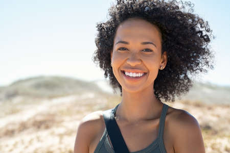 Portrait of young black woman with curly hair at beach. Smiling african american girl standing outdoor and looking at camera during summer vacation. Happy girl with bag at beach with copy space.の写真素材